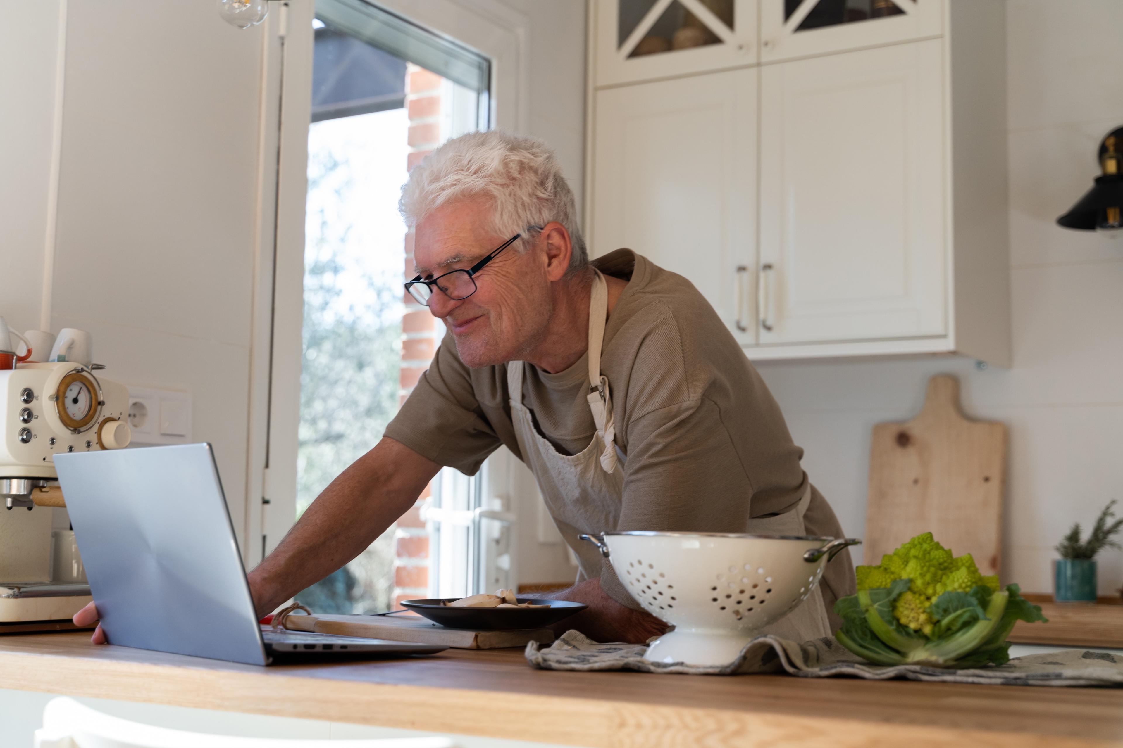 Homme d’âge moyen portant un tablier, debout près du comptoir de sa cuisine, entouré d’une variété d’ingrédients sains. Il sourit tout en regardant son ordinateur portable pendant un appel virtuel avec sa diététiste.